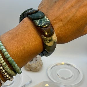 Könnte beinhalten: Ein klobiges Armband mit großen, rechteckigen Steinen in Grün-, Braun- und Cremetönen. Das Armband wird am Handgelenk getragen und zeigt die natürlichen Variationen in Farbe und Textur des Steins. Ein zweites Armband mit kleinen, runden Perlen ist ebenfalls sichtbar.