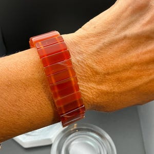 Könnte beinhalten: Ein elastisches Armband aus rechteckigen, polierten Karneolperlen in einem tiefen, durchscheinenden rot-orangen Farbton. Die Perlen sind in einer einzigen Reihe angeordnet und bilden ein durchgehendes Band, das glatt am Handgelenk anliegt.