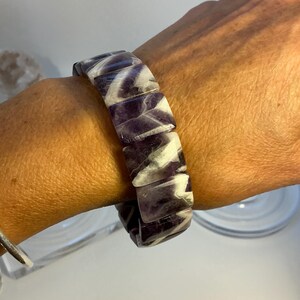 Puede incluir: Un brazalete grueso con piedras de amatista rectangulares con vetas blancas. El brazalete se lleva en la muñeca, mostrando el contraste de color púrpura y blanco. El diseño del brazalete es audaz y llamativo.