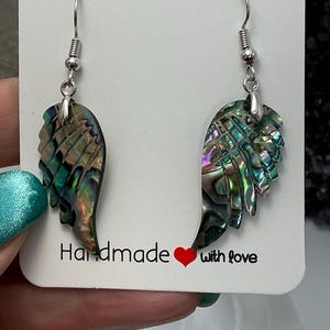 Puede incluir: Un par de pendientes de ala de concha de abul&oacute;n iridiscente hechos a mano. Los pendientes cuelgan de ganchos plateados. La tarjeta detr&aacute;s de los pendientes dice "Handmade with love."