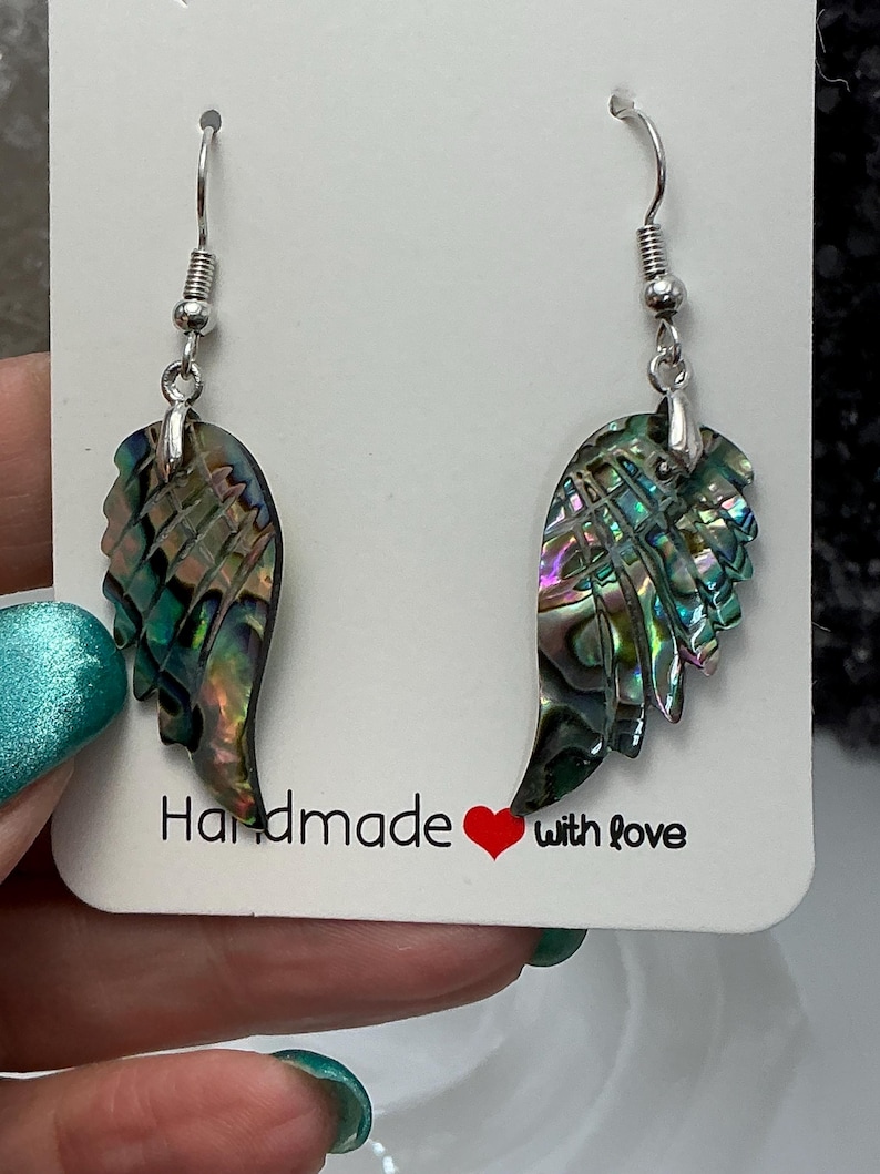 Puede incluir: Un par de pendientes de ala de concha de abul&oacute;n iridiscente hechos a mano. Los pendientes cuelgan de ganchos plateados. La tarjeta detr&aacute;s de los pendientes dice "Handmade with love."