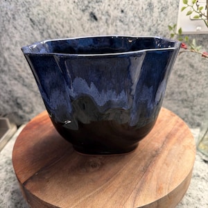 May include: A handcrafted ceramic bowl with a unique, wavy rim. The bowl features a gradient of dark blue to black glaze, creating a striking contrast. It sits on a round, wooden serving board, adding a rustic touch. This decorative bowl is ideal for serving or display.