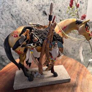 May include: A detailed figurine of a horse with an Native American theme. The horse is painted tan with red markings and a black mane and tail. It is adorned with a saddle, feathers, and a spear. The figurine is on a stone base.