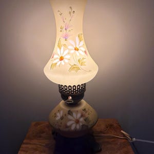 May include: A table lamp with a frosted glass shade and a matching glass base, both featuring hand-painted white daisy flowers with green leaves. The lamp has a dark metal burner-style collar with a small key switch and a dark base.