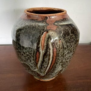 May include: A round ceramic vase with a wide opening and a tapered base. The exterior features a mottled glaze in shades of dark brown, charcoal, and cream, with exposed reddish-brown clay visible in vertical streaks and along the rim.