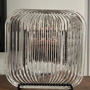 May include: Clear glass cloche with vertical ribbed design, resting on a black metal stand. The cloche has a square shape with rounded corners. The glass reflects the surrounding environment, creating a mirrored effect. The cloche is a decorative item.