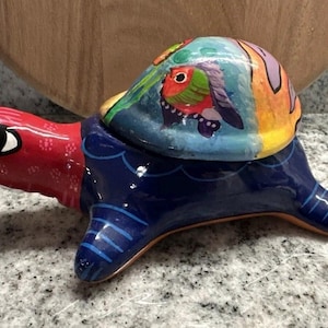 May include: A hand-painted ceramic turtle figurine. The turtle has a red head, a blue body, and a shell decorated with colorful bird and abstract designs. The figurine is a decorative item.