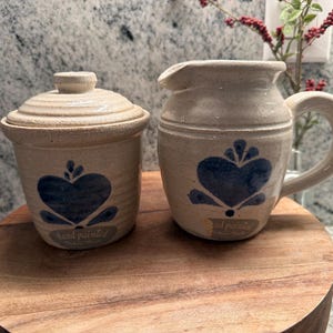 Puede incluir: Un azucarero de cerámica color crema con tapa y una jarra para crema con asa. Ambas piezas presentan un diseño de corazón azul y la inscripción "hand painted". El conjunto se exhibe sobre una superficie de madera.