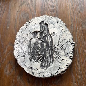 May include: A round, ceramic decorative plate with a detailed relief of a bird perched on a tree trunk. The bird and tree are dark gray, set against a cracked, off-white background. The plate has a rustic, handcrafted appearance.