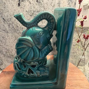 May include: Teal ceramic elephant bookend. The elephant head is detailed with a raised trunk and large ears. The bookend has a rectangular base and a decorative cutout design. The ceramic has a glossy finish.