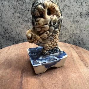 May include: A ceramic sculpture of a dragon head with a glossy finish in shades of cream, brown, and dark blue. The dragon head is mounted on a rectangular base with a marbled blue and white glaze, featuring detailed scales and facial features.