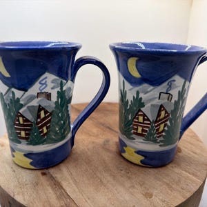 May include: Two ceramic mugs featuring a hand-painted winter scene with dark blue night skies, a yellow crescent moon, snow-covered mountains, green pine trees, and brown cabins with glowing yellow windows and chimneys emitting smoke.