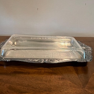 May include: A rectangular silver-toned metal butter dish featuring a clear glass lid. The base has an ornate, scalloped edge with embossed decorative detailing. The glass cover is transparent and smooth, designed to fit securely over the metal tray.