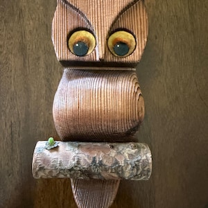 Pode incluir: Escultura de coruja em madeira com um design entalhado e texturizado. A coruja tem olhos grandes e redondos com detalhes amarelos e azuis. Está pousada num galho de madeira com um pequeno detalhe verde. Objeto decorativo.