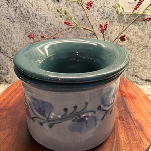 May include: A ceramic butter dish with a teal lid and a floral design in blue and gray. The dish is cylindrical and sits on a wooden surface. The floral design wraps around the body of the dish.