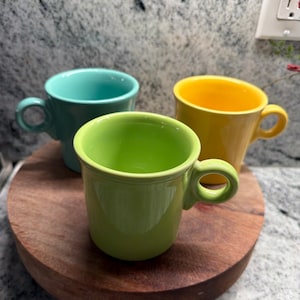 Op de afbeelding: Drie keramische mokken in heldere effen kleuren: één limoengroen, één turquoise en één geel. Elke mok heeft een gladde, glanzende afwerking, een cilindrisch lichaam en een afgerond handvat aan de zijkant. De mokken staan bij elkaar opgesteld.