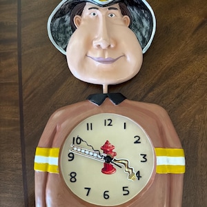 May include: A novelty wall clock shaped like a smiling firefighter. The clock face features a fire hydrant and ladder design. The firefighter's hat reads "Fireman". The clock is brown, beige, black, yellow, and red.