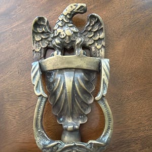 May include: A brass door knocker featuring a detailed eagle perched atop a central shield-like element with a scalloped design. The lower portion consists of a curved, ornate handle that connects to the base, showcasing a weathered metallic finish.