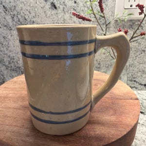 May include: A tall, cylindrical ceramic mug with a glossy tan finish and speckled texture. The mug features two sets of double horizontal blue stripes, one set near the top rim and one set near the base, and includes a sturdy, angled handle on the side.