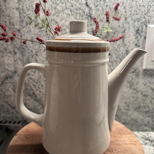 May include: Cream-colored ceramic coffee pot with a brown stripe around the lid. The pot has a handle and a spout. It is sitting on a wooden surface. The background includes a sprig of red berries.