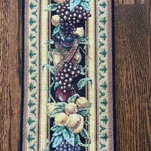 May include: A tapestry runner with a fruit design, including grapes, plums, and pears, on a black background. It has a cream border with a floral pattern and wooden dowels at the top. The runner is suitable for a table.