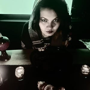 May include: A dimly lit scene featuring a person in a dark hooded garment, gazing intently. A decorative glass orb, candles in small jars, and a patterned dish are on a dark surface, creating a mystical ambiance.