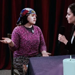 May include: A woman wearing a patterned headscarf and a pink sweater sits across from another woman holding a microphone. A lit candle and a small glass are on the table between them. The background features red curtains.