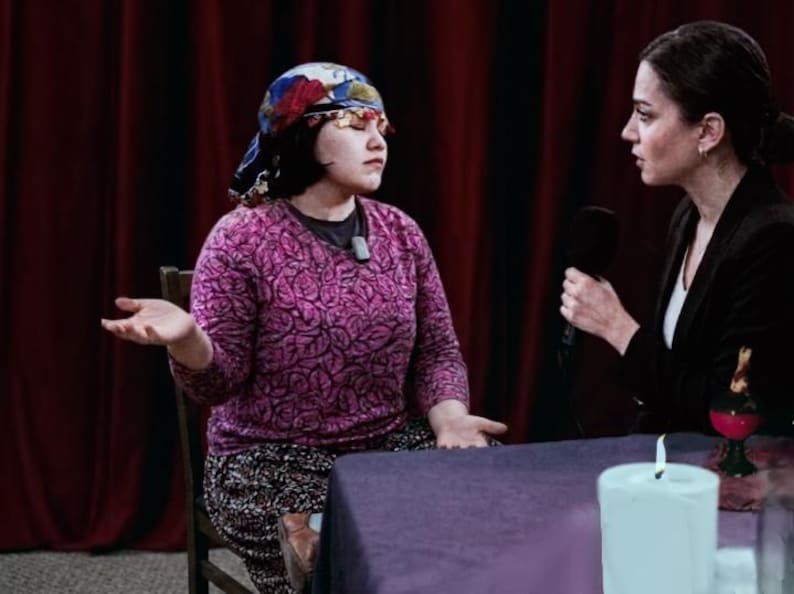 May include: A woman wearing a patterned headscarf and a pink sweater sits across from another woman holding a microphone. A lit candle and a small glass are on the table between them. The background features red curtains.