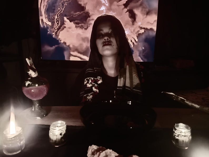 May include: A person in a dark setting with candles and a potion bottle. The person is wearing a dark outfit with floral embroidery. A screen in the background displays a cloudy, colorful image. There are also small jars with candles.
