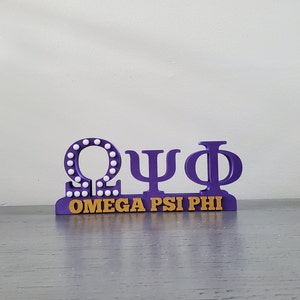 Puede incluir: Letras griegas Omega Psi Phi moradas con puntos blancos alrededor de la letra Omega y letras doradas debajo que dicen "OMEGA PSI PHI".