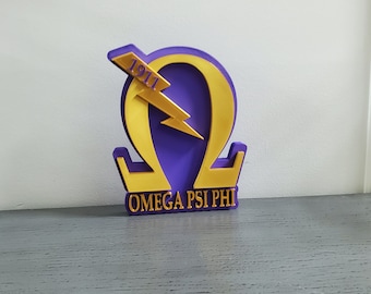 May include: A purple and gold 3D printed fraternity symbol with the Greek letter Omega and a lightning bolt. The symbol has the year 1911 and the words "OMEGA PSI PHI" printed on it.