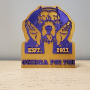 以下が含まれることがあります： 金と紫色の3Dプリントされた犬の頭部の置物で、台座に「EST. 1911 OMEGA PSI PHI」の文字が書かれています。