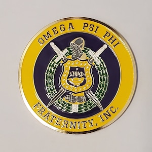 May include: A gold-coloured metal coin with a purple and green emblem featuring a shield with the letters "ΩΨΦ" and two crossed swords. The coin has the text "OMEGA PSI PHI FRATERNITY, INC." around the edge.