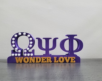 May include: Purple and gold Greek letters "Omega Psi Phi" with white dots around the Omega. The letters are on a purple base with the words "Wonder Love" in gold letters.