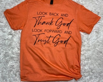 Pode incluir: Camiseta laranja com texto preto que diz "Olhe para trás e agradeça a Deus, olhe para frente e confie em Deus."