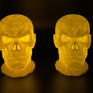May include: Two glowing skull-shaped lamps with a yellow light. The lamps have a detailed design, with visible eye sockets, a nose cavity, and teeth. The background is black, which makes the lamps stand out. These lamps are ideal for Halloween or gothic decor.