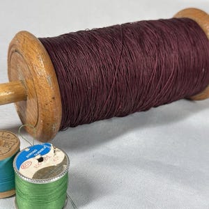 May include: A large wooden spool of dark burgundy thread with a wooden handle, and two smaller spools of thread in teal and green. The large spool is the focal point, suggesting a sewing or craft theme.