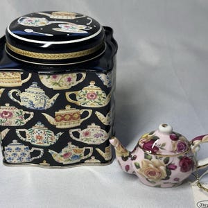 May include: A square black metal tea tin featuring a repeating pattern of various colorful teapots and a matching lid, accompanied by a small, round, light pink ceramic teapot decorated with a floral rose pattern and gold-tone trim.