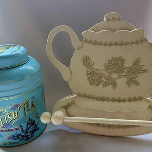 May include: A light blue tin of Traditional English Tea with gold accents and a decorative teapot image. Beside it is a cream-colored teapot-shaped paper towel holder with floral detailing and a wooden dowel.