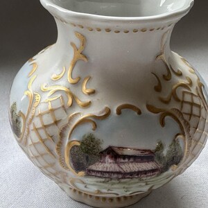 May include: A small, off-white ceramic vase with a landscape scene and gold accents. The vase features a painted image of a building with a red roof and trees. Gold filigree-style designs adorn the vase's surface.