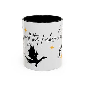 Pode incluir: Caneca de cerâmica branca com interior preto, apresentando uma silhueta de dragão preto, estrelas e a frase "well the fuck away" em escrita preta. Acentuada com explosões de estrelas douradas.