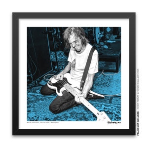 K&ouml;nnte beinhalten: Ein Schwarzwei&szlig;foto einer Person, die Bassgitarre spielt. Die Person sitzt auf einem blauen, gemusterten Teppich. Das Foto ist in einem schwarzen Rahmen mit einem wei&szlig;en Passepartout. Der Text "Malamuth of today 30 (2017)" und "ROBXWALLACE" ist auf dem Bild zu sehen.