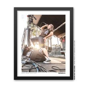 May include: A framed photograph captures a live music performance. A person is singing into a microphone on stage, with stage equipment and a crowd in the background. The image is in color, with the artist wearing a dark shirt and shorts.