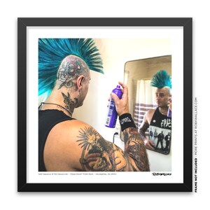 May include: A man with a blue mohawk and tattoos sprays hairspray on his hair while looking in the mirror. The mirror reflects his back and the text "ROBXWALLACE" is visible in the bottom right corner of the mirror.