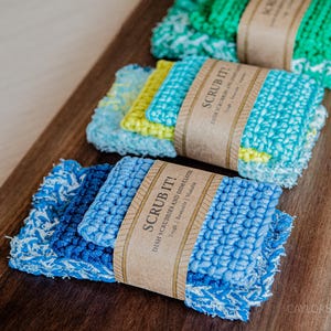 May include: A wooden cutting board displays three stacks of colorful dish scrubbies. Each stack is wrapped with a brown paper band that reads "SCRUB IT!" in bold letters. The scrubbies are in shades of blue, green, and yellow.
