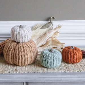 May include: Three knitted pumpkins in different colors and sizes. The largest pumpkin is brown, the middle pumpkin is white, and the smallest pumpkin is blue. The pumpkins are sitting on a woven rug.