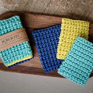 May include: Four crocheted dish scrubbers in blue, yellow, and turquoise. The scrubbers are stacked on a wooden cutting board. The turquoise scrubber has a label that reads "Scrub It! Dish Scrubbers | Reusable | Washable."