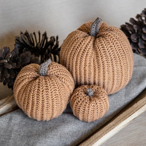 May include: Three knitted pumpkins in various sizes, brown with a ribbed texture, sitting on a gray fabric surface in a wooden tray.