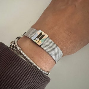 May include: A silver mesh bracelet with a silver charm featuring the BTS logo and the letters "BTS" in black.