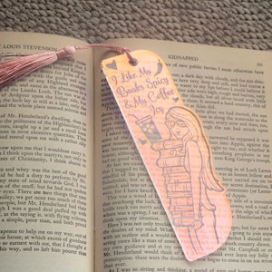 Peut inclure: Un marque-page rose et brun avec un gland. Le marque-page présente une illustration de dessin animé d'une femme tenant une pile de livres et une tasse de café. Le texte sur le marque-page est "J'aime mes livres épicés et mon café joyeux".